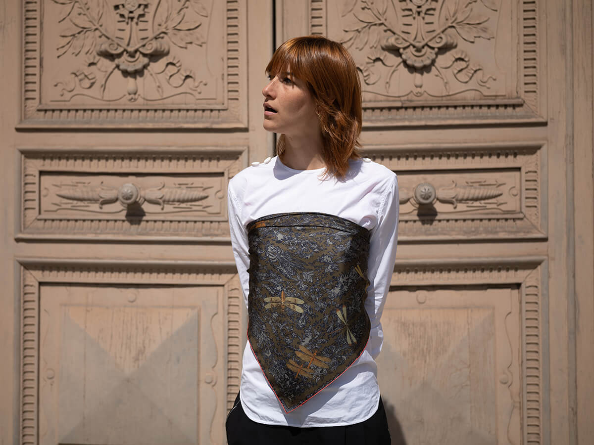 young lady wearing a white shirt and a black silk scarf with dragonfly motif around her chest with a door made of wood in the background