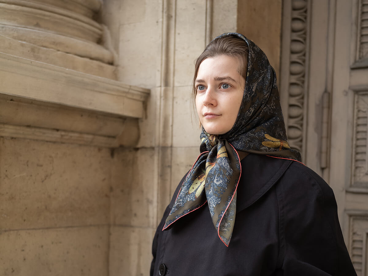 jeune femme portant un foulard en soie bleu avec motif libellule autour de la tête et un manteau noir avec une porte en bois en arrière plan